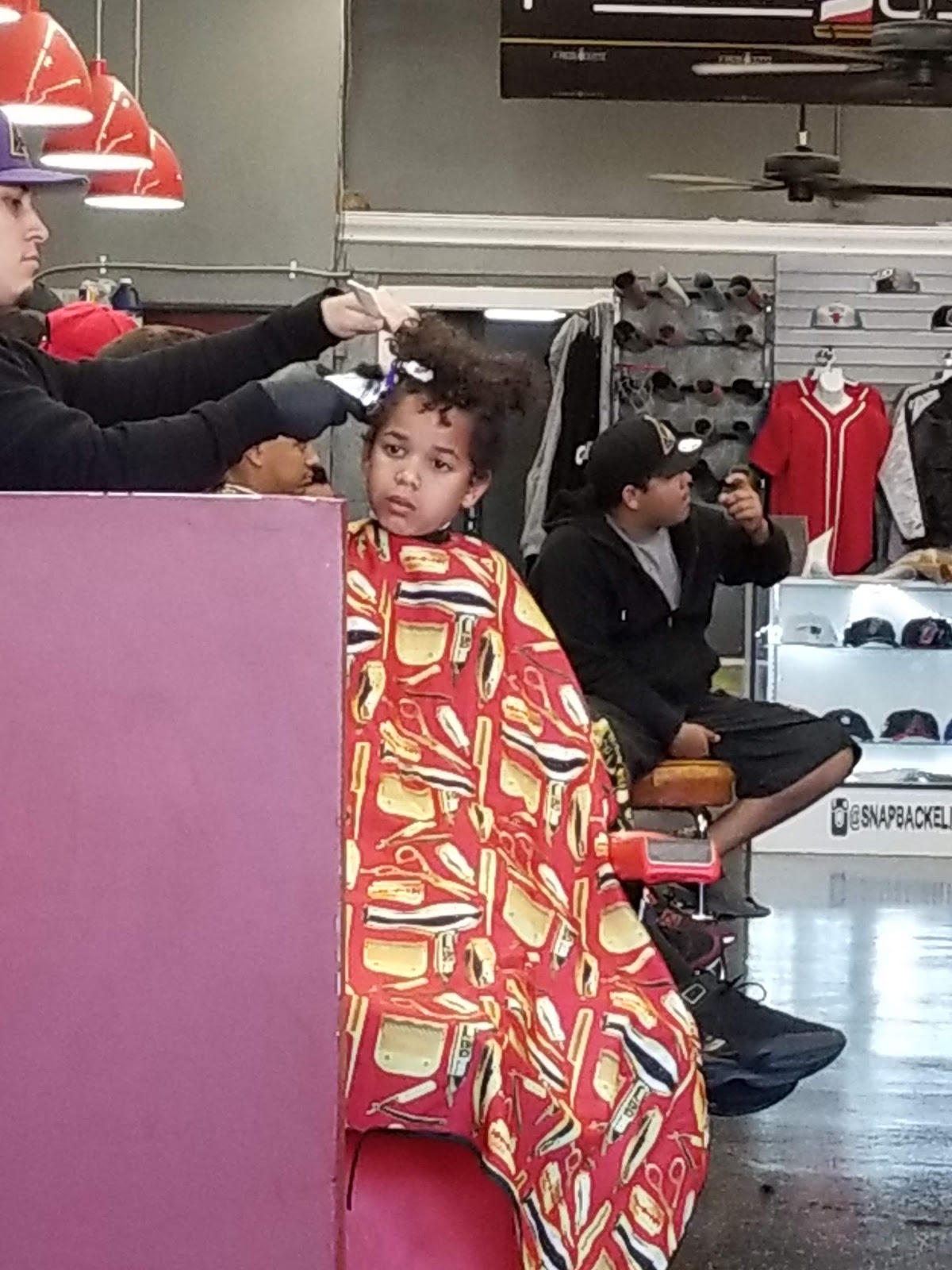 A young customer getting his curls shaped up in a red tool-print cape, with our snapback wall and jersey case behind him